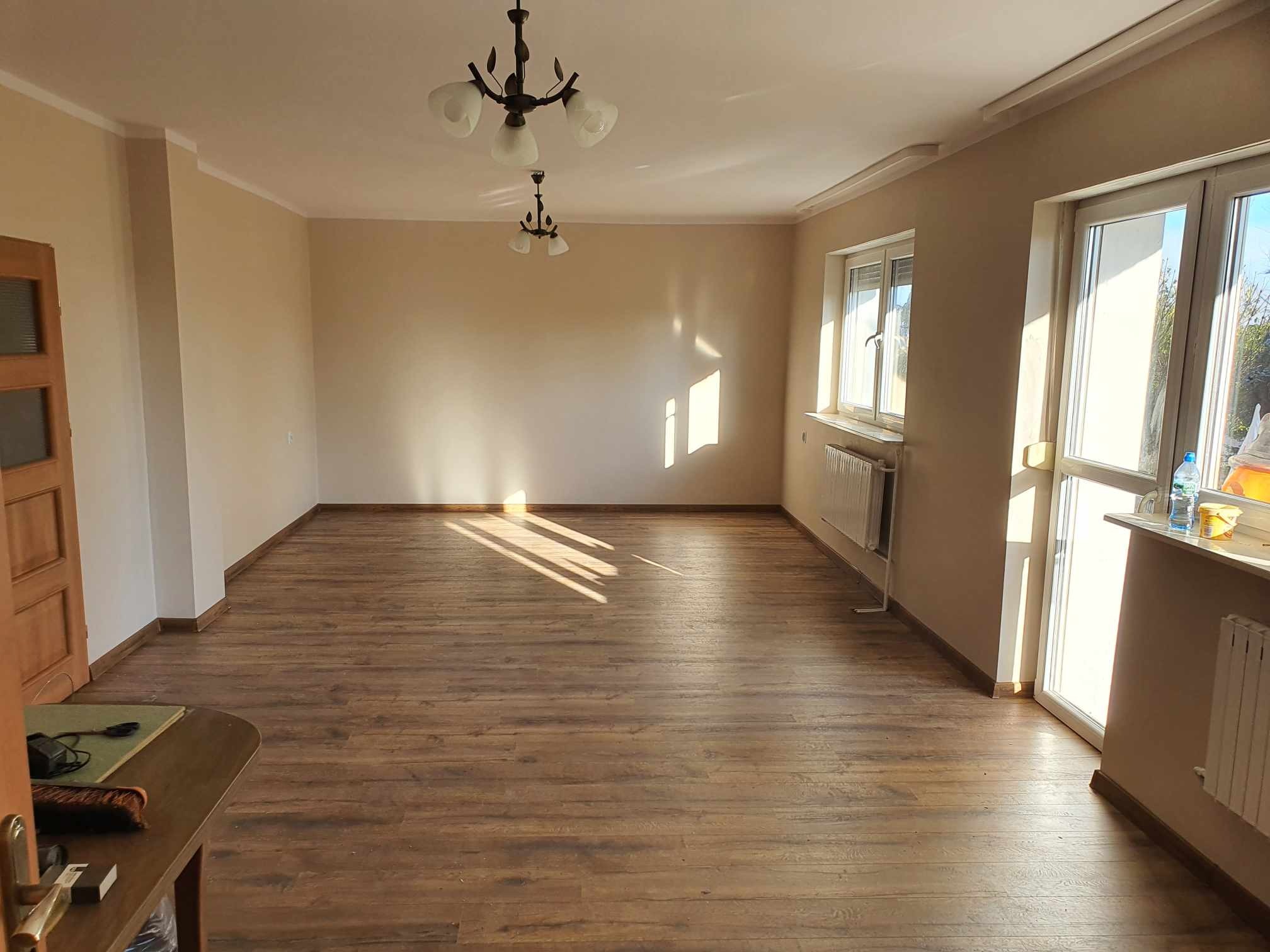 Pusty pokój po remoncie z drewnianą podłogą, beżowymi ścianami, białymi oknami i dwiema lampami sufitowymi w stylu retro, zalany popołudniowym światłem.