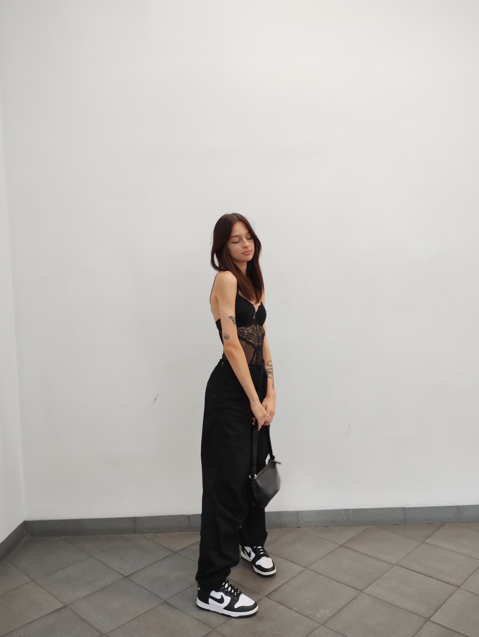 Portret kobiety w czarnym koronkowym body i szerokich spodniach, pozującej na tle białej ściany w Bydgoszczy, z torebką na ramieniu i tatuażami na ramionach.