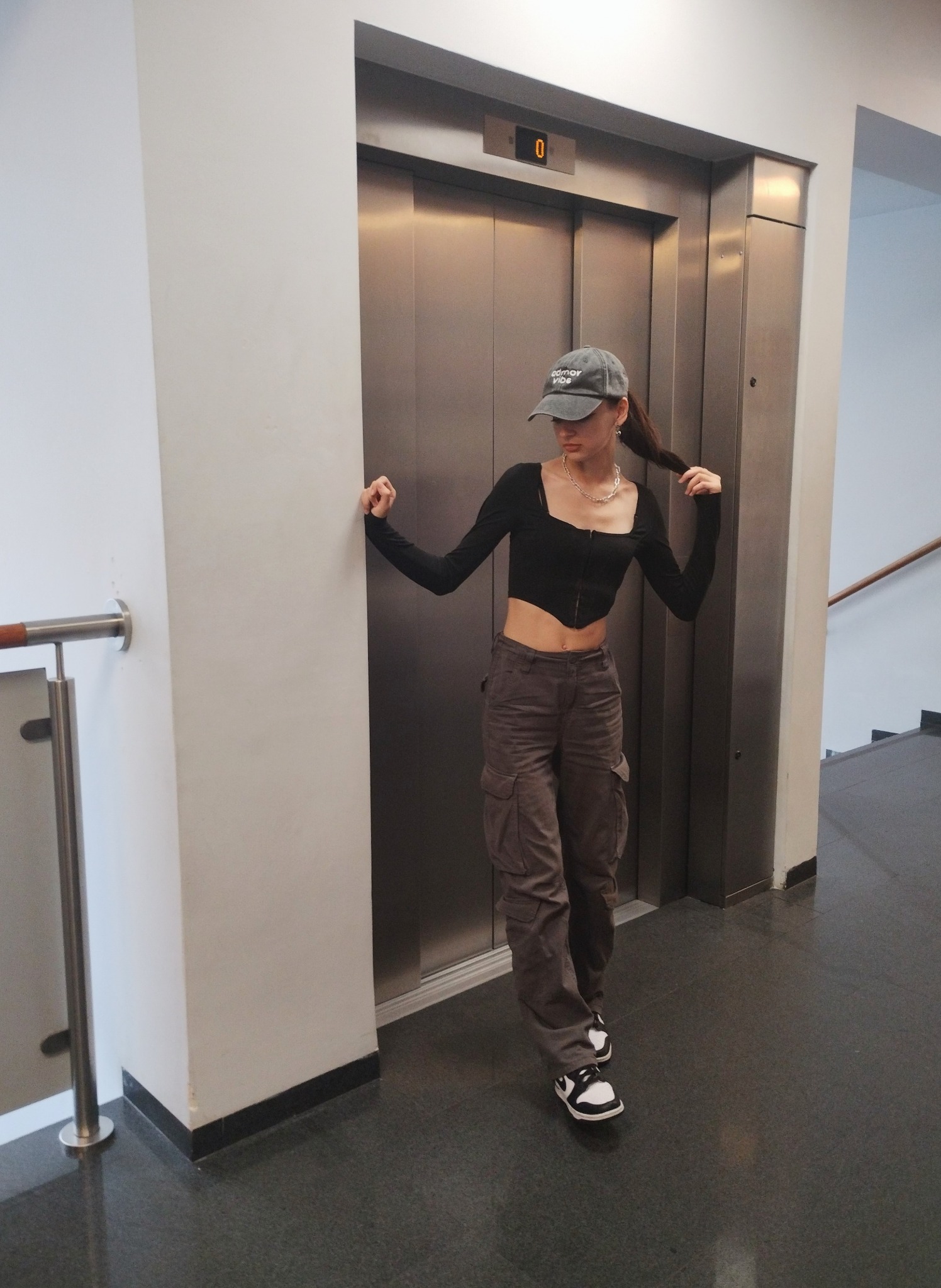 Młoda kobieta w czarnym topie i szarych spodniach cargo pozuje obok metalowych drzwi windy, trzymając włosy w dłoni.