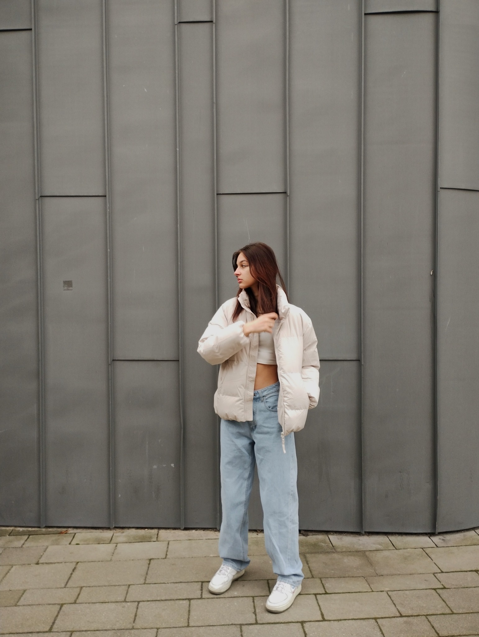 Młoda kobieta w jasnej kurtce puchowej, topie i szerokich jeansach pozuje na tle szarej, metalowej ściany z pionowymi panelami.