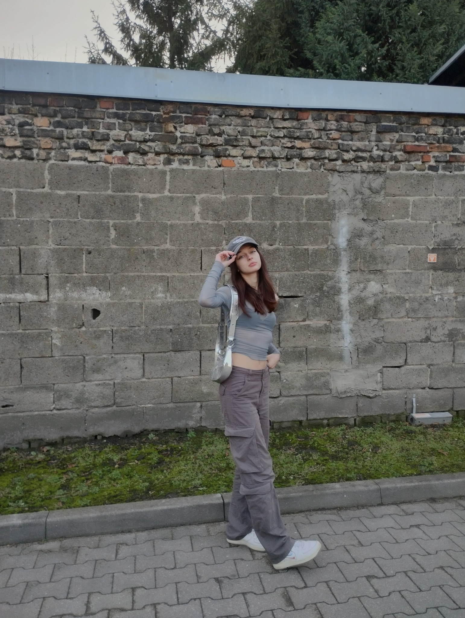 Portret młodej kobiety w szarej bluzce i spodniach cargo, pozującej na tle szarego, ceglanego muru z mchem i zielenią u podstawy.