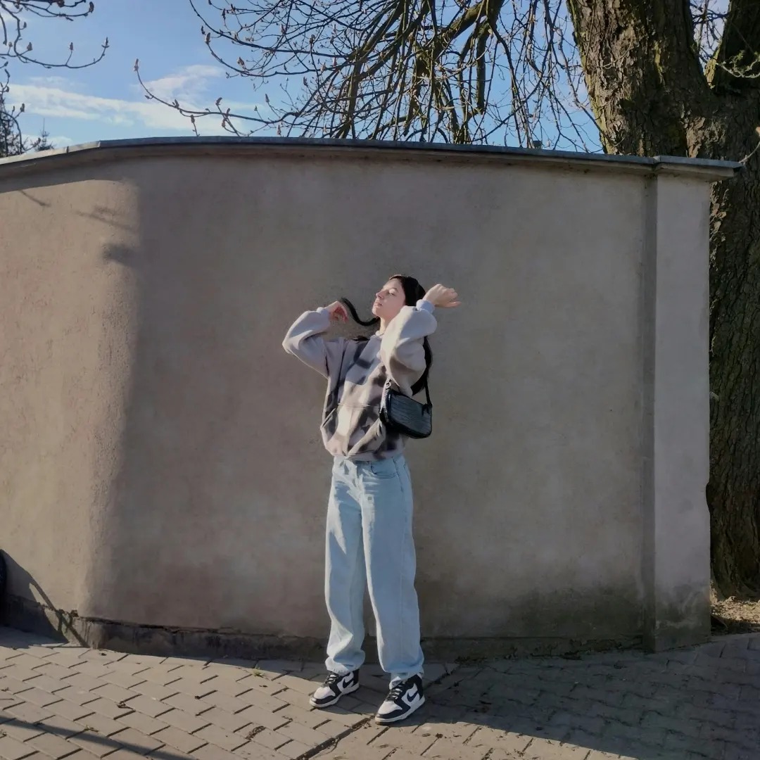 Młoda kobieta w bluzie tie-dye i szerokich jeansach pozuje na tle betonowego muru w słoneczny dzień, z rękami uniesionymi nad głową i małą czarną torebką na ramieniu.