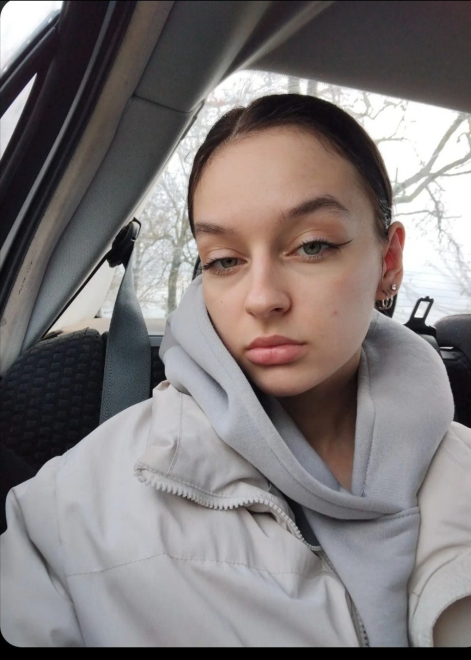 Portret młodej kobiety z delikatnym makijażem oczu, ubrana w bluzę z kapturem i kurtkę, siedzącej w samochodzie z widocznym pasem bezpieczeństwa.