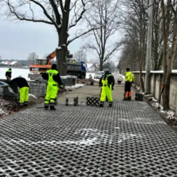 Pracownicy w odblaskowych ubraniach montują ażurową nawierzchnię z betonowych elementów, w tle wywrotka i koparka CAT, zimowa sceneria z resztkami śniegu.