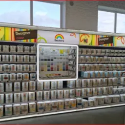 Ekspozycja puszek z farbami marki Beckers Designer Colour na regale sklepowym, z paletą kolorów umieszczoną w centralnej części. Widoczne okna w tle i fragment ekspozycji baterii kuchennych po...