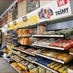 Wnętrze sklepu budowlanego z regałami pełnymi narzędzi malarskich, taśm, pędzli i innych akcesoriów. W tle ekspozycja paneli imitujących kamień.