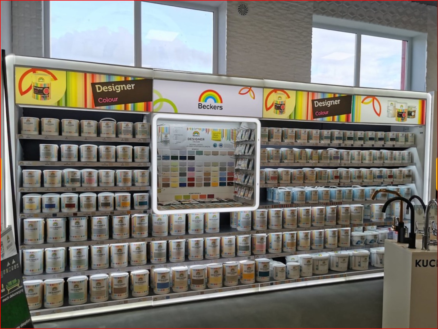 Ekspozycja puszek z farbami marki Beckers Designer Colour na regale sklepowym, z paletą kolorów umieszczoną w centralnej części. Widoczne okna w tle i fragment ekspozycji baterii kuchennych po...