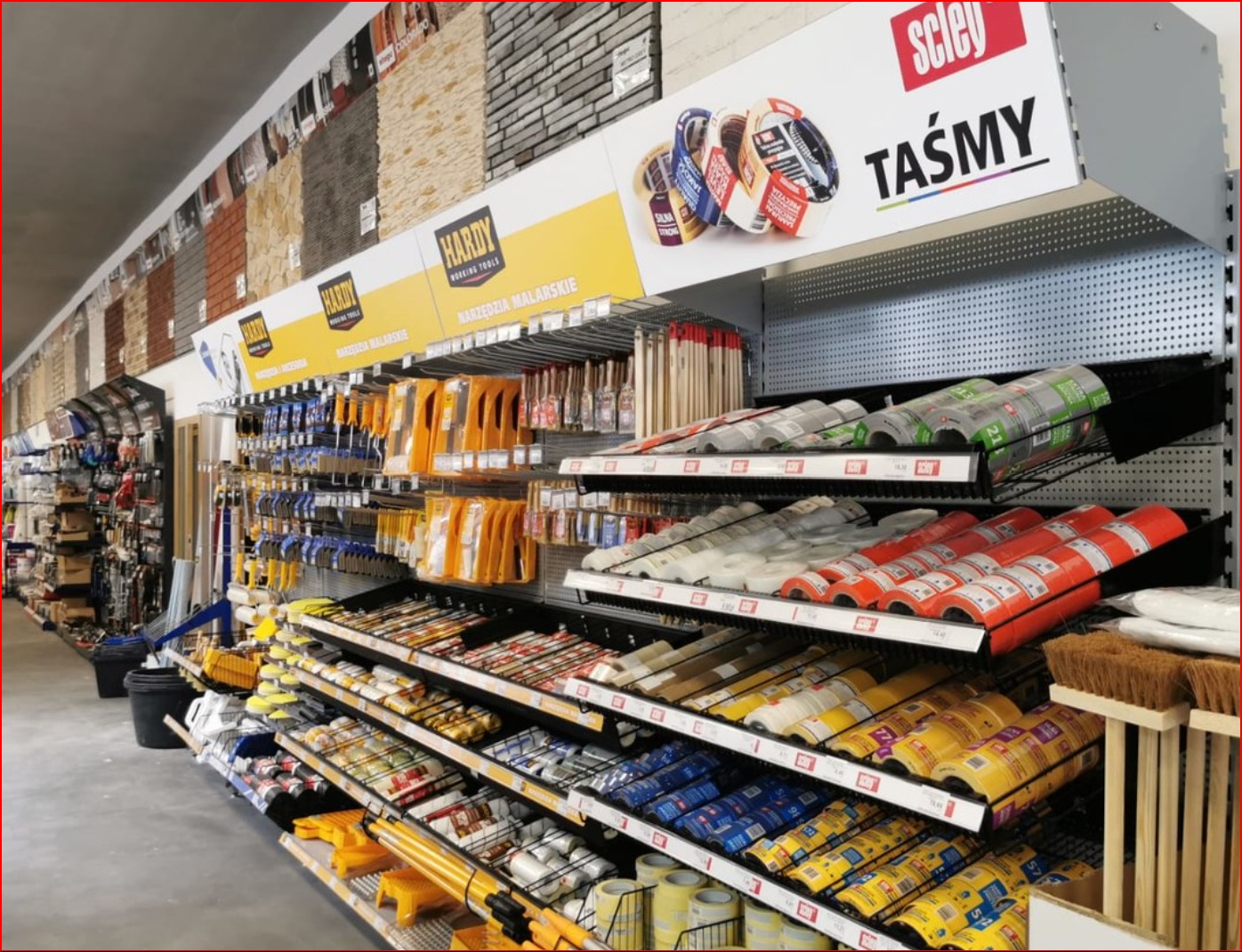 Wnętrze sklepu budowlanego z regałami pełnymi narzędzi malarskich, taśm, pędzli i innych akcesoriów. W tle ekspozycja paneli imitujących kamień.