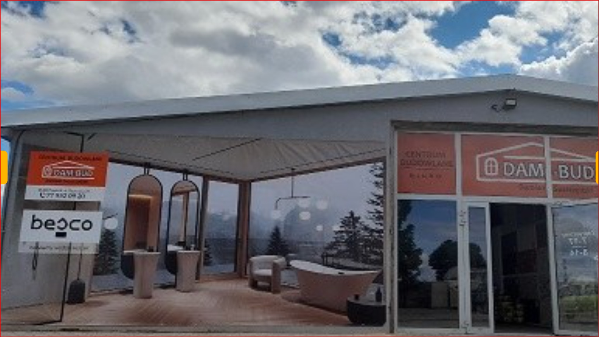 Ekspozycja aranżacji łazienki wewnątrz namiotu handlowego, z widocznym logo 'DAM-BUD' oraz 'beco' i numerem telefonu, na tle górskiego krajobrazu.