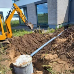 Żółta mini koparka wykopuje rów pod szarą rurę kanalizacyjną przy fundamencie nowoczesnego domu jednorodzinnego, widoczna szara studzienka kanalizacyjna z betonową pokrywą, dwie łopaty wbite w ziemię.