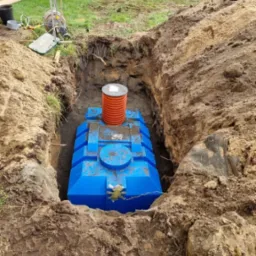 Niebieski, plastikowy zbiornik na szambo umieszczony w wykopie ziemnym, z widocznym kominkiem wentylacyjnym i śladami opon ciężkiego sprzętu na ziemi.