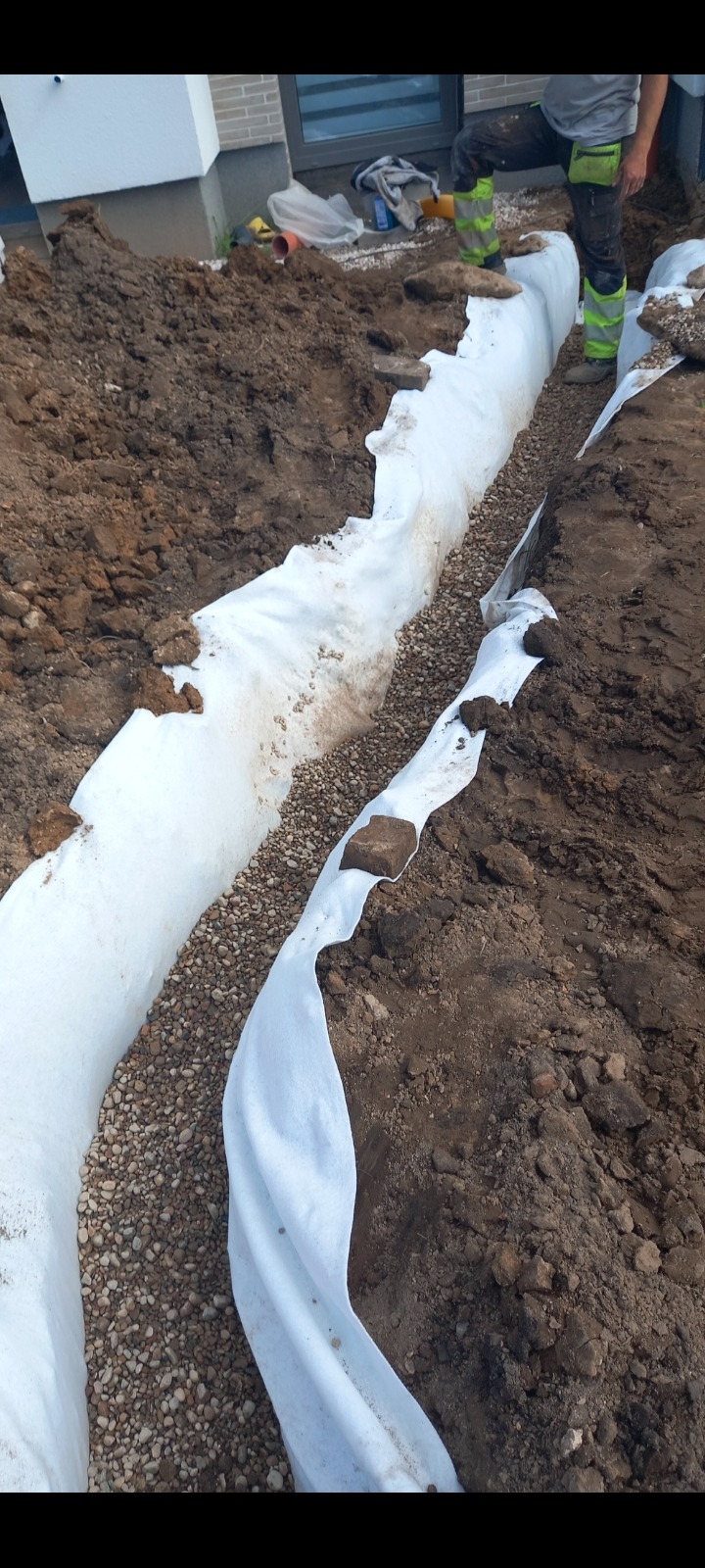 Wykop liniowy wyłożony białą geowłókniną, wypełniony drobnym żwirem, widoczny fragment budynku i pracownik w odblaskowych butach.