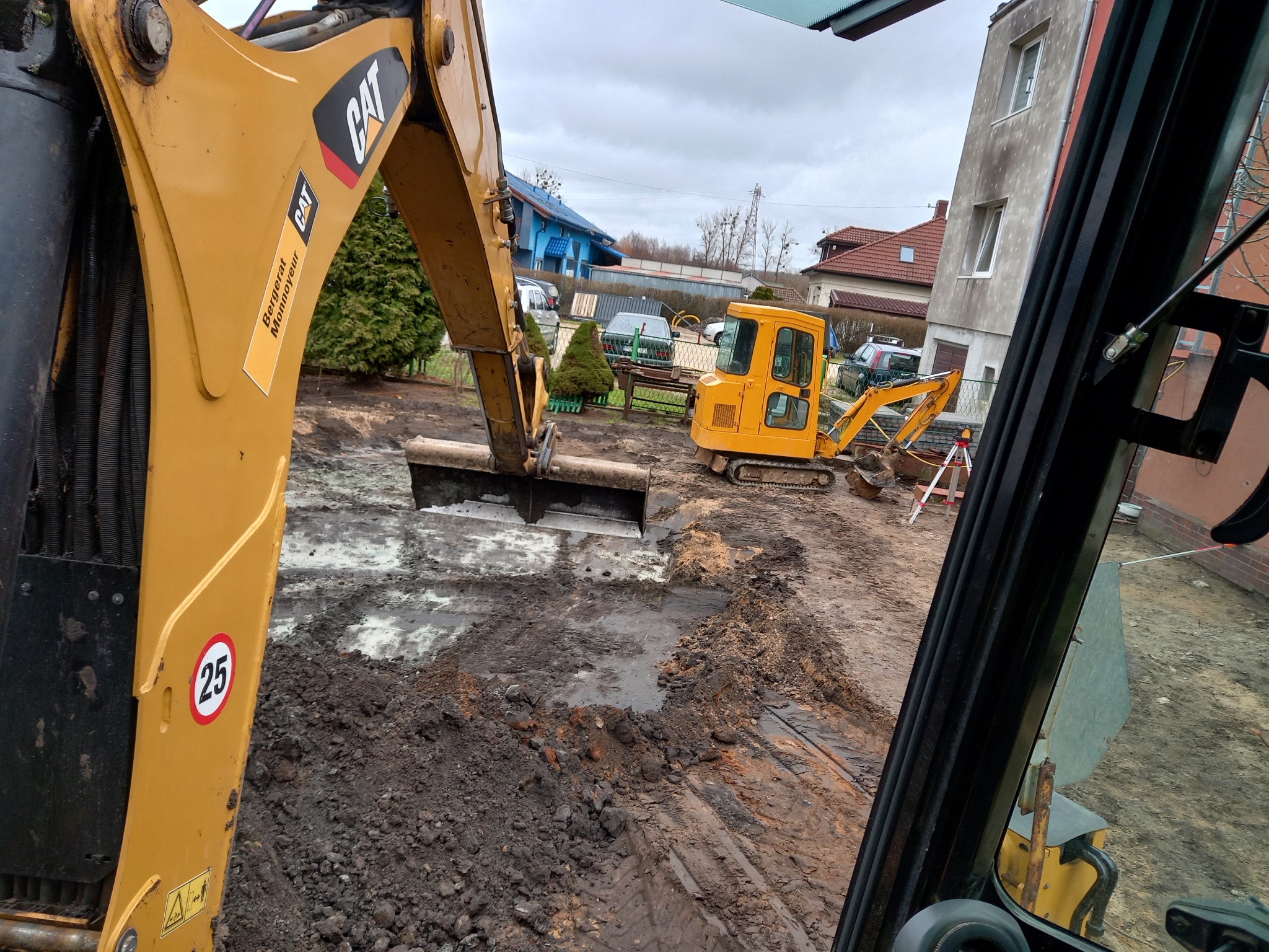 Żółty minikoparka CAT z łyżką w trakcie prac ziemnych na działce, widok z kabiny innego sprzętu, w tle domy jednorodzinne i pochmurne niebo.