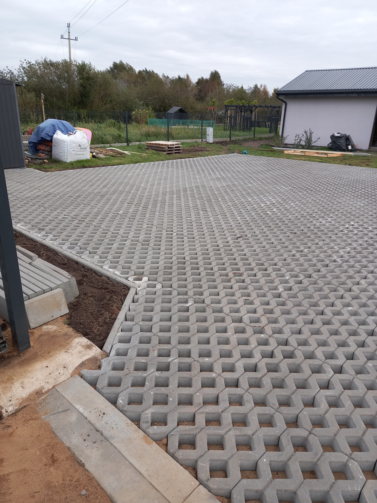 Ułożona szara ażurowa kostka brukowa na podjeździe, widoczne worki z materiałami budowlanymi i przygotowane obrzeża, w tle dom i zieleń.