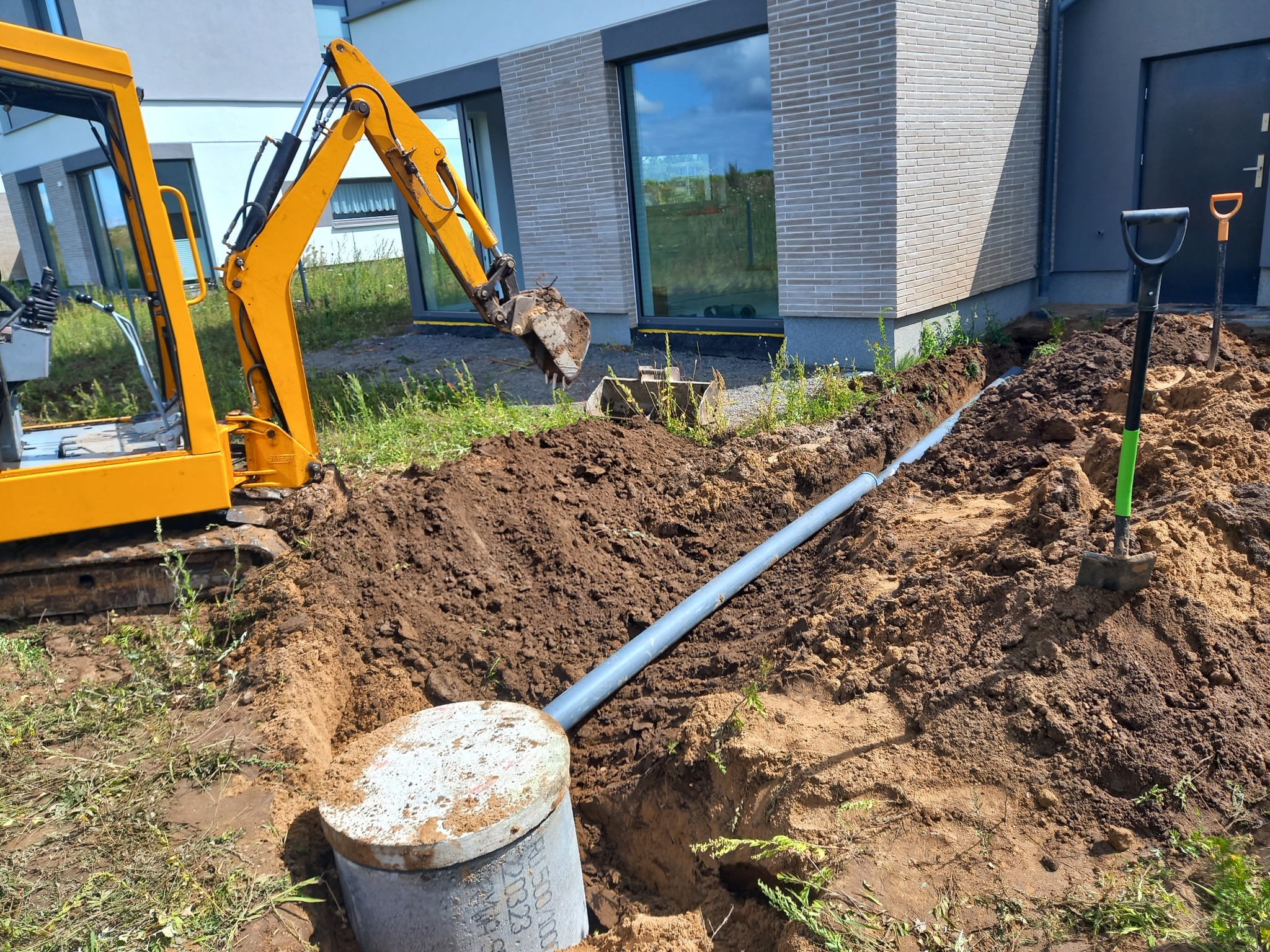 Żółta mini koparka wykopuje rów pod szarą rurę kanalizacyjną przy fundamencie nowoczesnego domu jednorodzinnego, widoczna szara studzienka kanalizacyjna z betonową pokrywą, dwie łopaty wbite w ziemię.