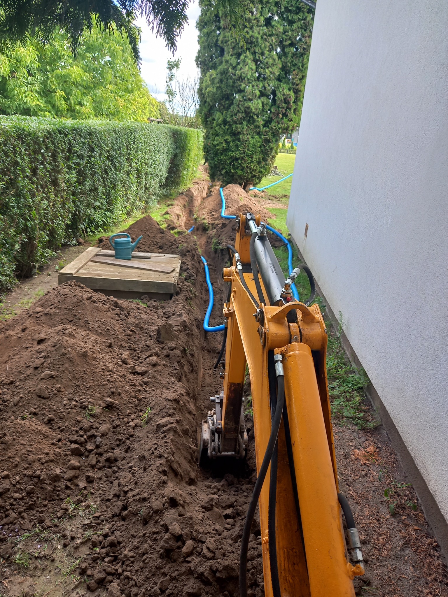 Żółta minikoparka w trakcie wykonywania wykopu pod niebieskie rury wzdłuż ściany budynku i żywopłotu; na pierwszym planie sterta ziemi i drewniana studzienka z zielonym konewką.
