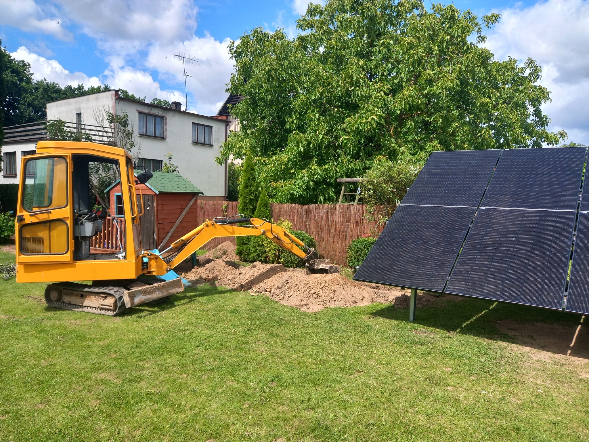 Żółta mini koparka przygotowuje teren pod montaż paneli fotowoltaicznych w ogrodzie, widoczne panele, trawa, dom i drzewa w tle.