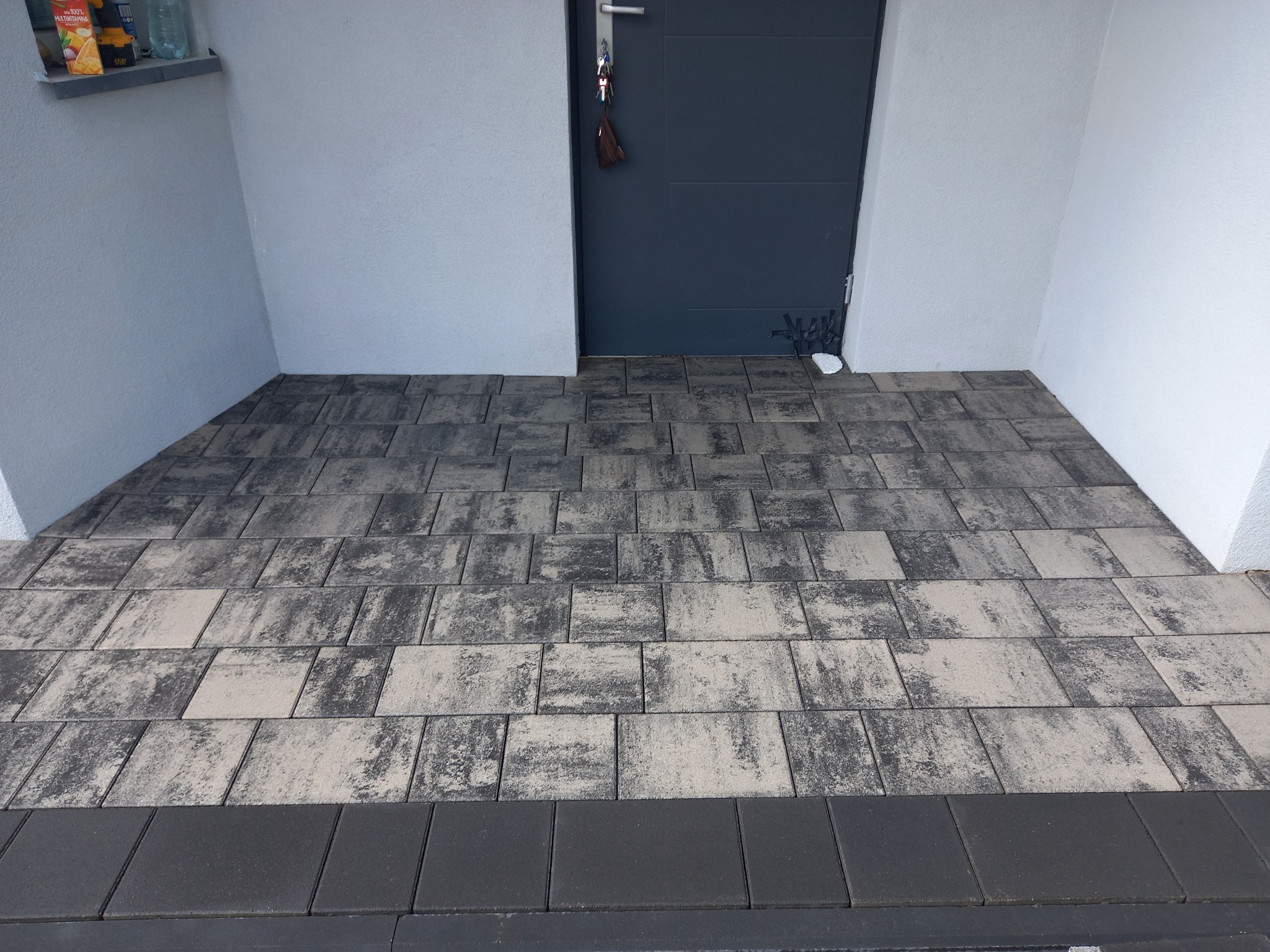 Ułożona, szara kostka brukowa przed wejściem do budynku z szarą, metalową furtką i białymi ścianami. Na furtce wiszą klucze.