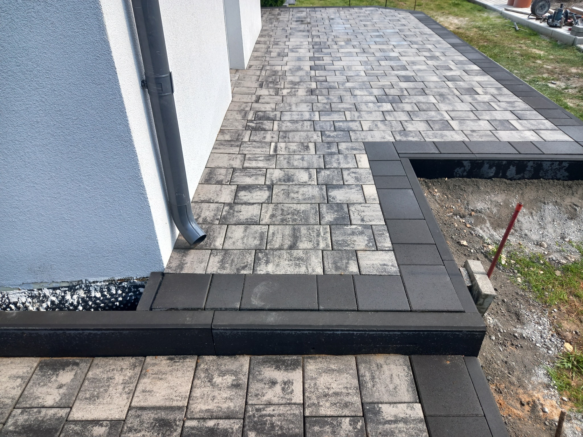 Ułożona kostka brukowa dwukolorowa (szaro-beżowa i grafitowa) z obrzeżem, widoczna rynna spustowa przy ścianie budynku i przygotowane podłoże z boku