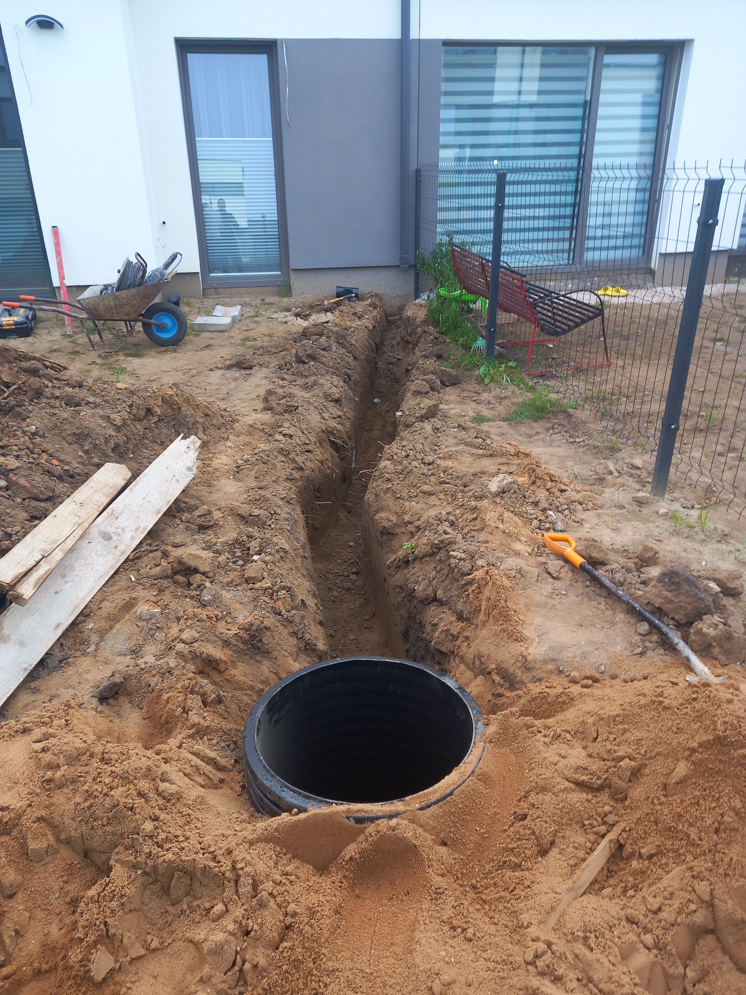 Czarna studzienka kanalizacyjna w wykopie obok nowoczesnego domu z szarą elewacją, widoczna łopata i taczka z narzędziami.