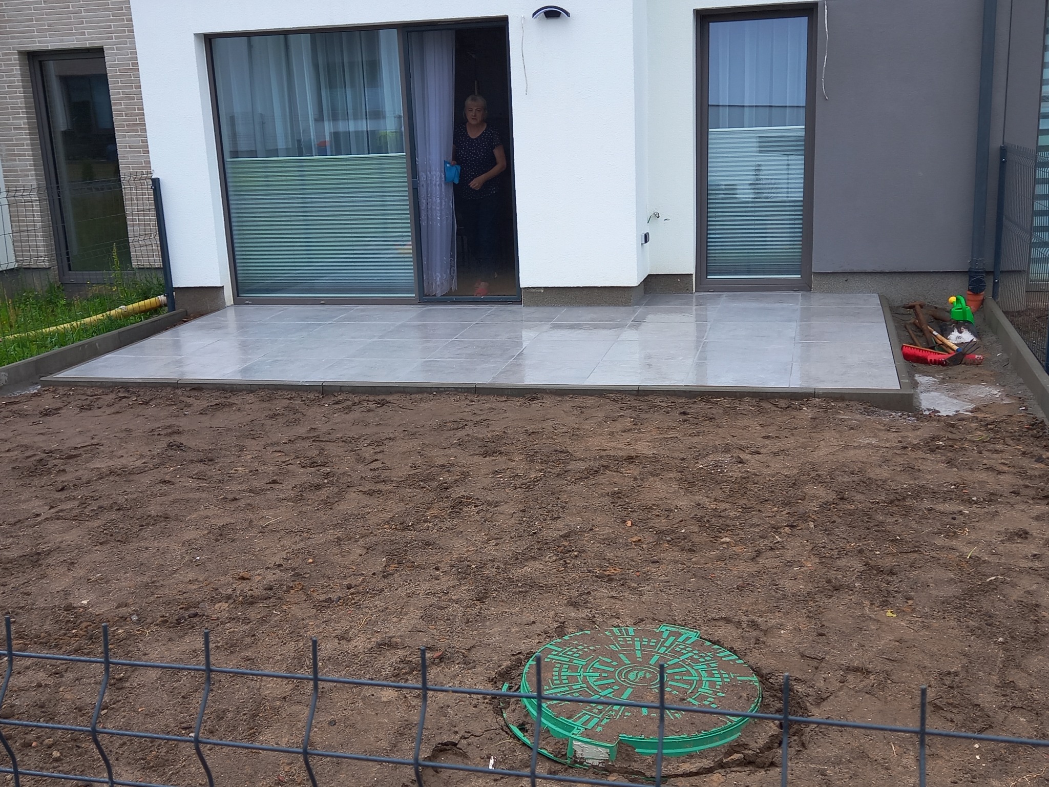 Nowoczesny taras z szarych płytek ceramicznych, widok z ogrodu na dom z przesuwnymi drzwiami, kobieta w drzwiach, przygotowany teren pod dalsze prace ziemne, zielona studzienka kanalizacyjna.