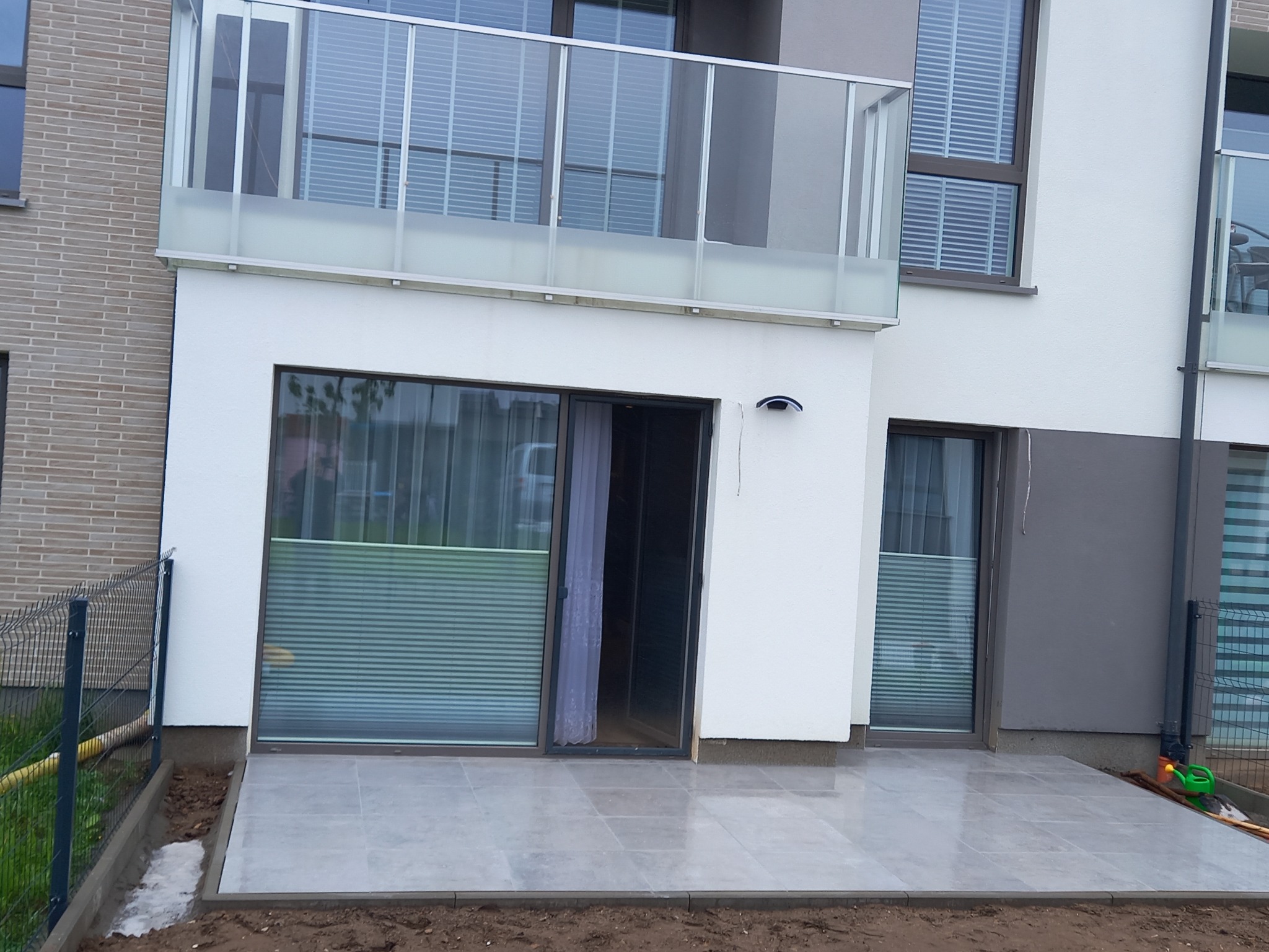 Nowoczesny taras wyłożony jasnoszarą płytką, z widokiem na budynek z balkonami i siatką ogrodzeniową w tle. Widoczny fragment ziemi przygotowany pod dalsze prace.