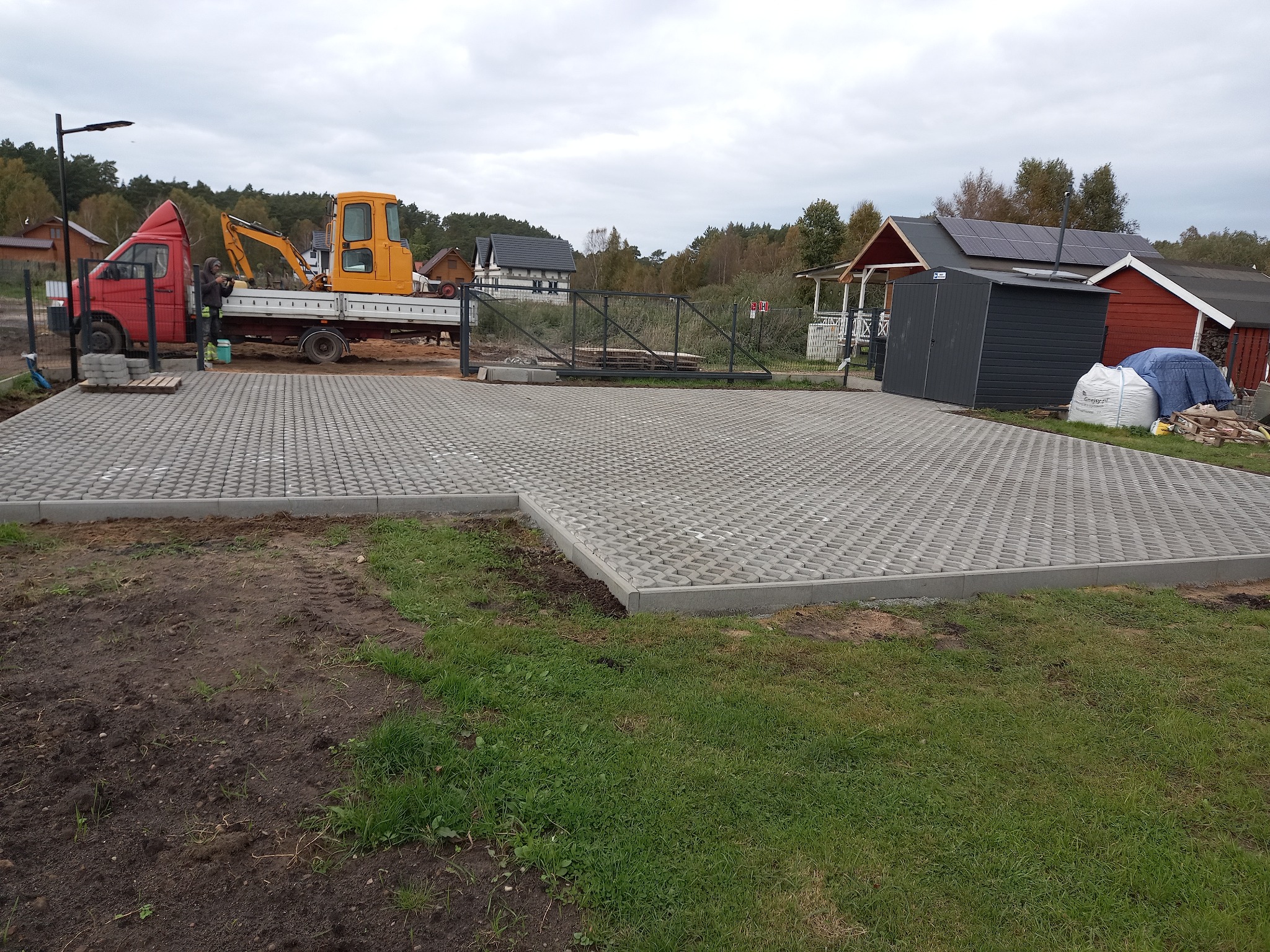 Ukończona nawierzchnia z ażurowych betonowych płyt, obrzeża z kostki, na podwórku stoi czerwono-biały samochód dostawczy z mini koparką na pace, w tle zabudowania gospodarcze.