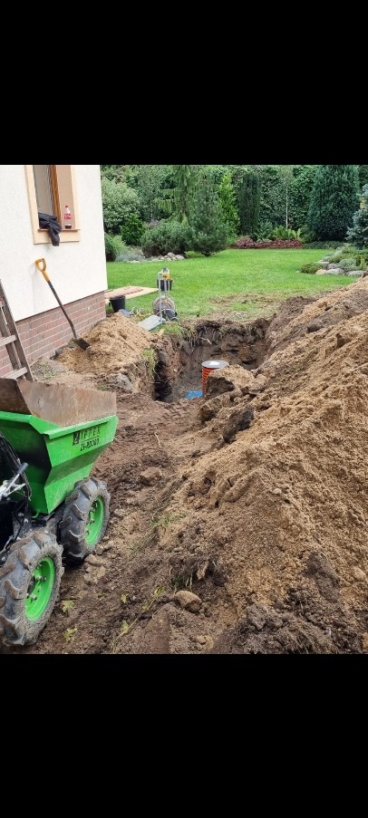 Wykop pod instalacje w ogrodzie, widoczny zielony wozidło budowlane, sterta ziemi, szpadel oparty o ścianę budynku, w tle trawnik i drzewa.