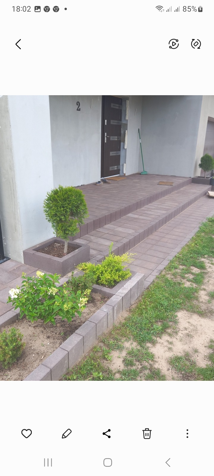 Schody wejściowe do domu wyłożone brązową kostką brukową, z donicami z roślinami po bokach i numerem domu '2' nad drzwiami.