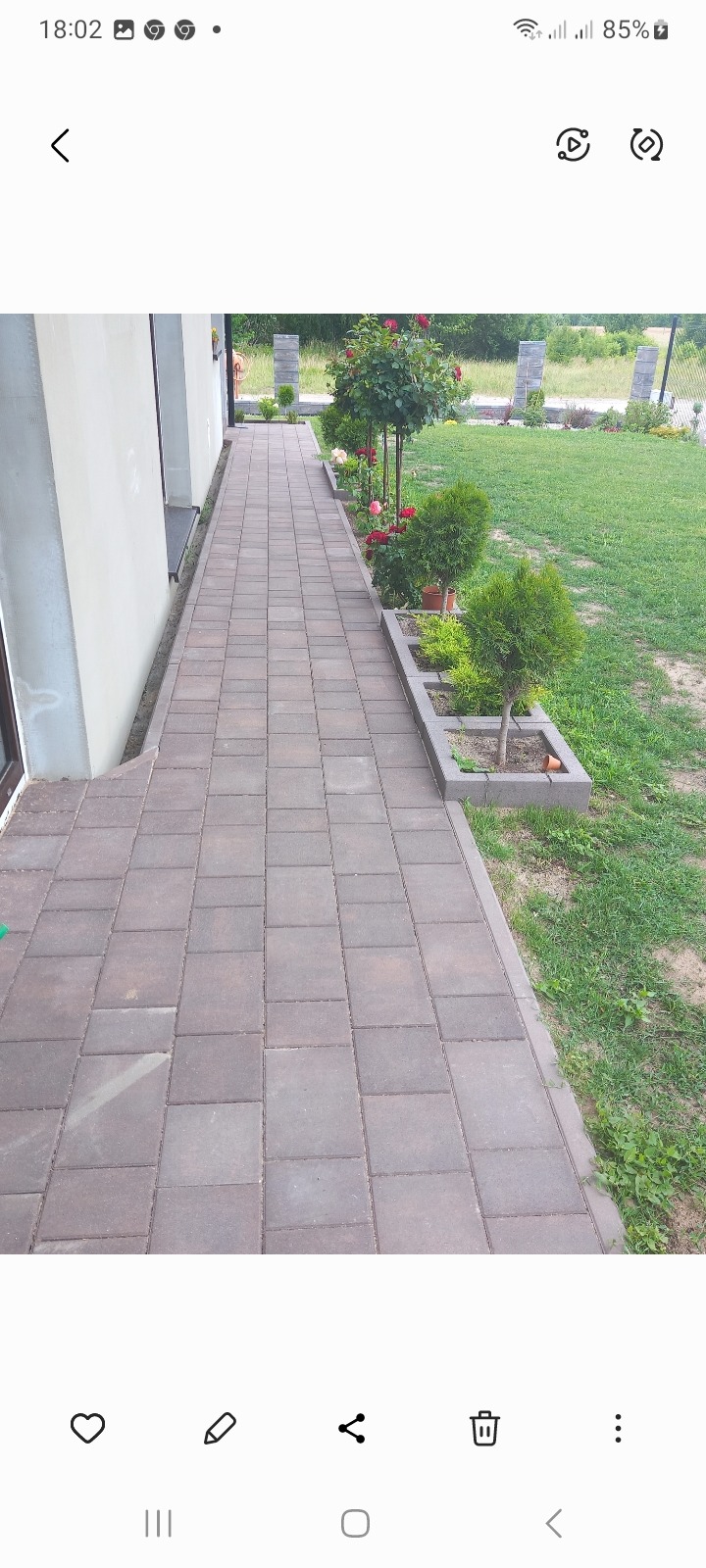 Ułożona ścieżka z kostki brukowej prowadząca wzdłuż budynku, z rabatami kwiatowymi i krzewami po prawej stronie, widok na trawnik i ogrodzenie w tle.