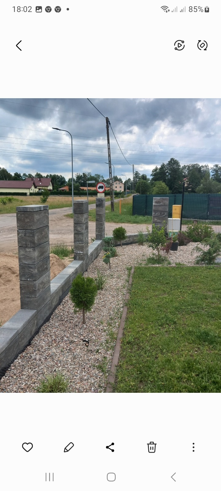 Nowoczesny, szary murek z betonowych bloczków z filarami, oddzielający posesję od drogi z zakazem wjazdu ciężarówek, wysypany drobnymi kamykami i obsadzony młodymi roślinami.