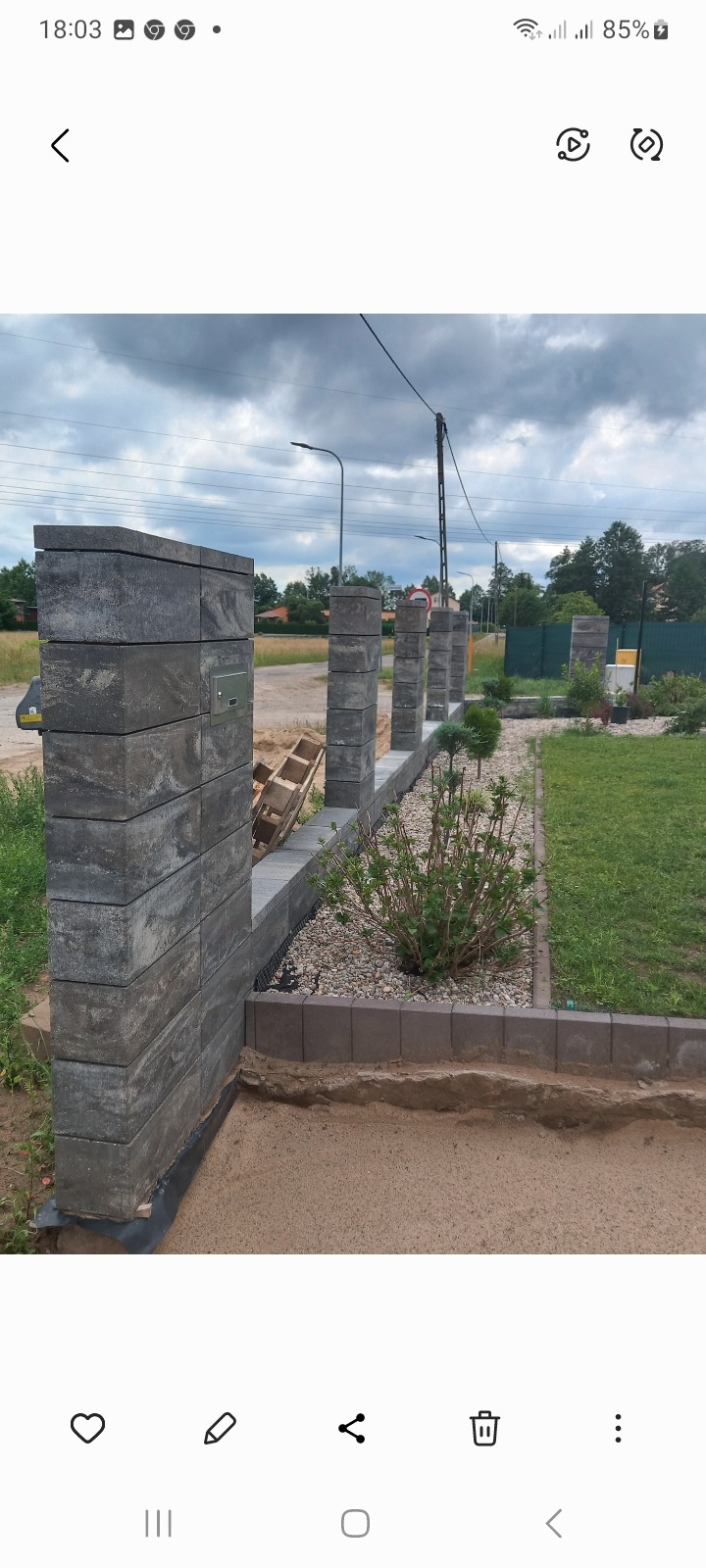 Nowo wybudowane ogrodzenie z szarych, betonowych bloczków z zamontowaną skrzynką na listy, obok rabata z krzewami i wysypanym kamieniem ozdobnym.