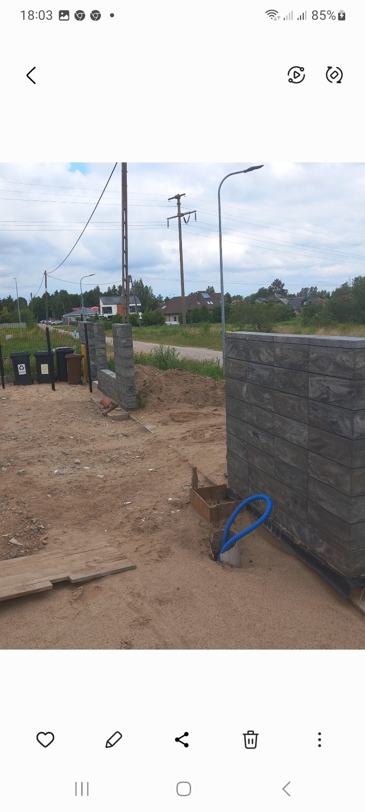 Wznoszony mur z szarych bloczków betonowych na piaszczystym podłożu, widoczne słupki ogrodzeniowe i rury instalacyjne, w tle domy i drzewa.