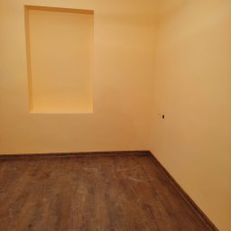 LEPARD - Pomalowane na żółto wnętrze pokoju z ułożonymi panelami podłogowymi, widoczna wnęka w ścianie i listwa dekoracyjna pod sufitem, mały otwór w ścianie