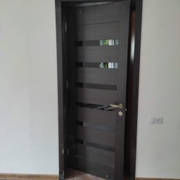 LEPARD - Ciemne, nowoczesne drzwi wewnętrzne z poziomymi przeszkleniami, uchylone w świeżo wyremontowanym pomieszczeniu z białymi ścianami i panelami podłogowymi imitującymi drewno. Widoczny kontakt...