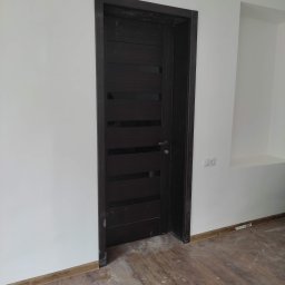 LEPARD - Ciemne, nowoczesne drzwi wewnętrzne z poziomymi przeszkleniami zamontowane w białej ścianie z dekoracyjnym gzymsem sufitowym i widocznymi śladami prac wykończeniowych na podłodze z paneli.