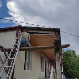 LEPARD - Pracownik na kolanach na rusztowaniu, montuje drewniane deski pod zadaszeniem domu jednorodzinnego. Widoczna drabina oparta o ścianę budynku, obok stoi starsza kobieta.