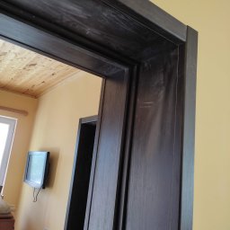 LEPARD - Ciemna, drewniana ościeżnica drzwiowa z widocznymi śladami użytkowania i drobnymi zarysowaniami, na tle żółtej ściany i drewnianego sufitu.