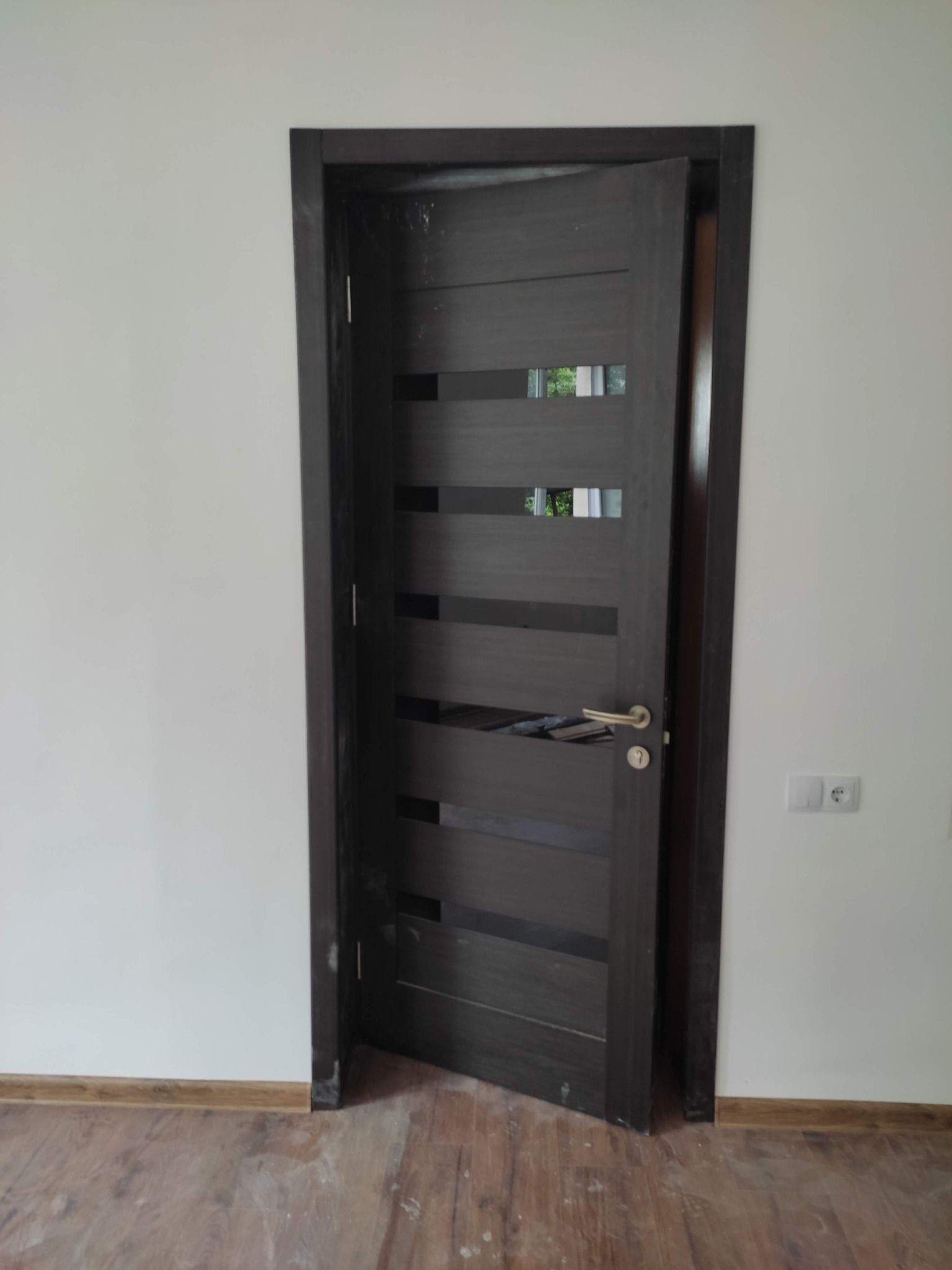 Ciemne, nowoczesne drzwi wewnętrzne z poziomymi przeszkleniami, uchylone w świeżo wyremontowanym pomieszczeniu z białymi ścianami i panelami podłogowymi imitującymi drewno. Widoczny kontakt...