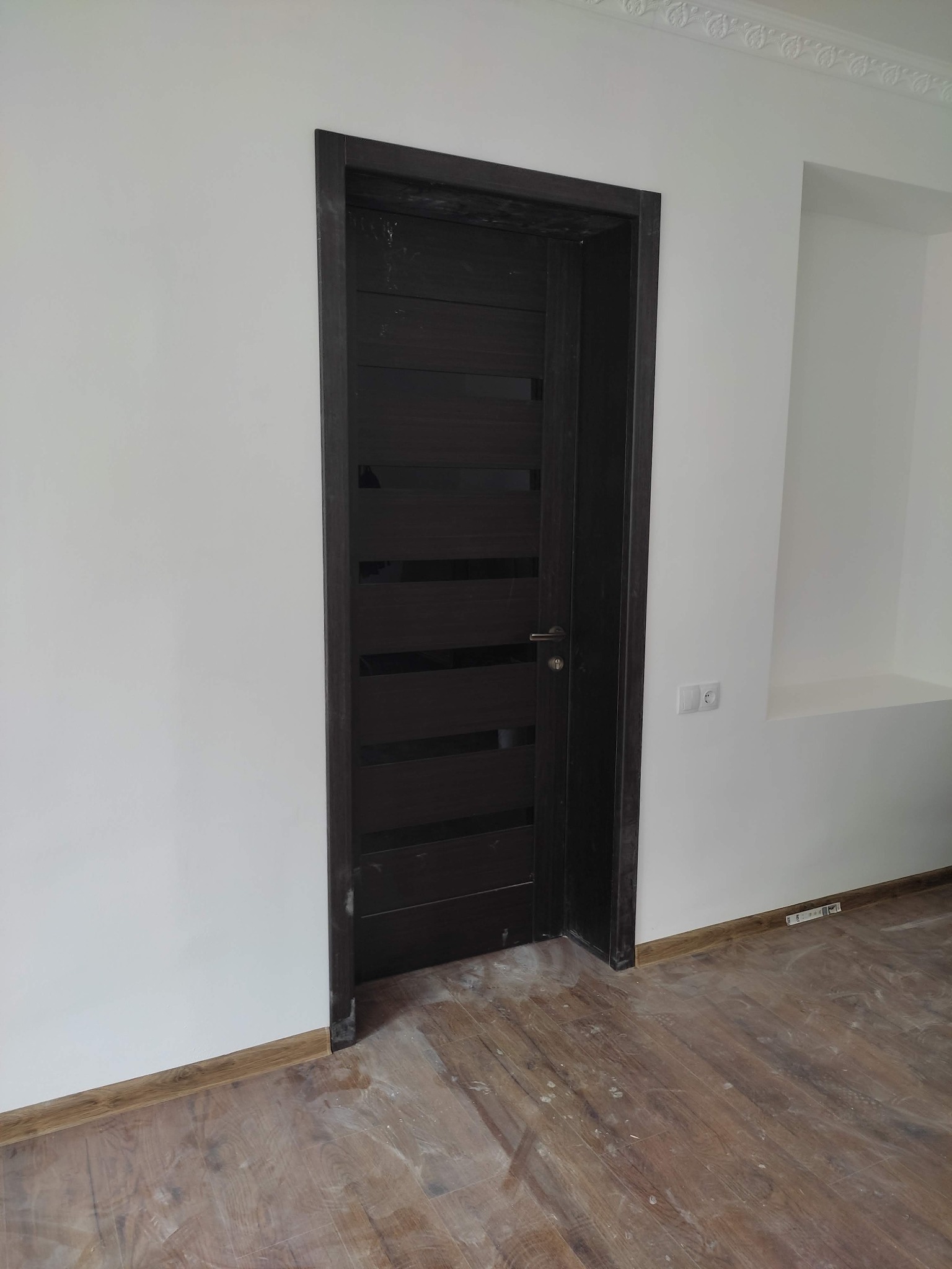 Ciemne, nowoczesne drzwi wewnętrzne z poziomymi przeszkleniami zamontowane w białej ścianie z dekoracyjnym gzymsem sufitowym i widocznymi śladami prac wykończeniowych na podłodze z paneli.