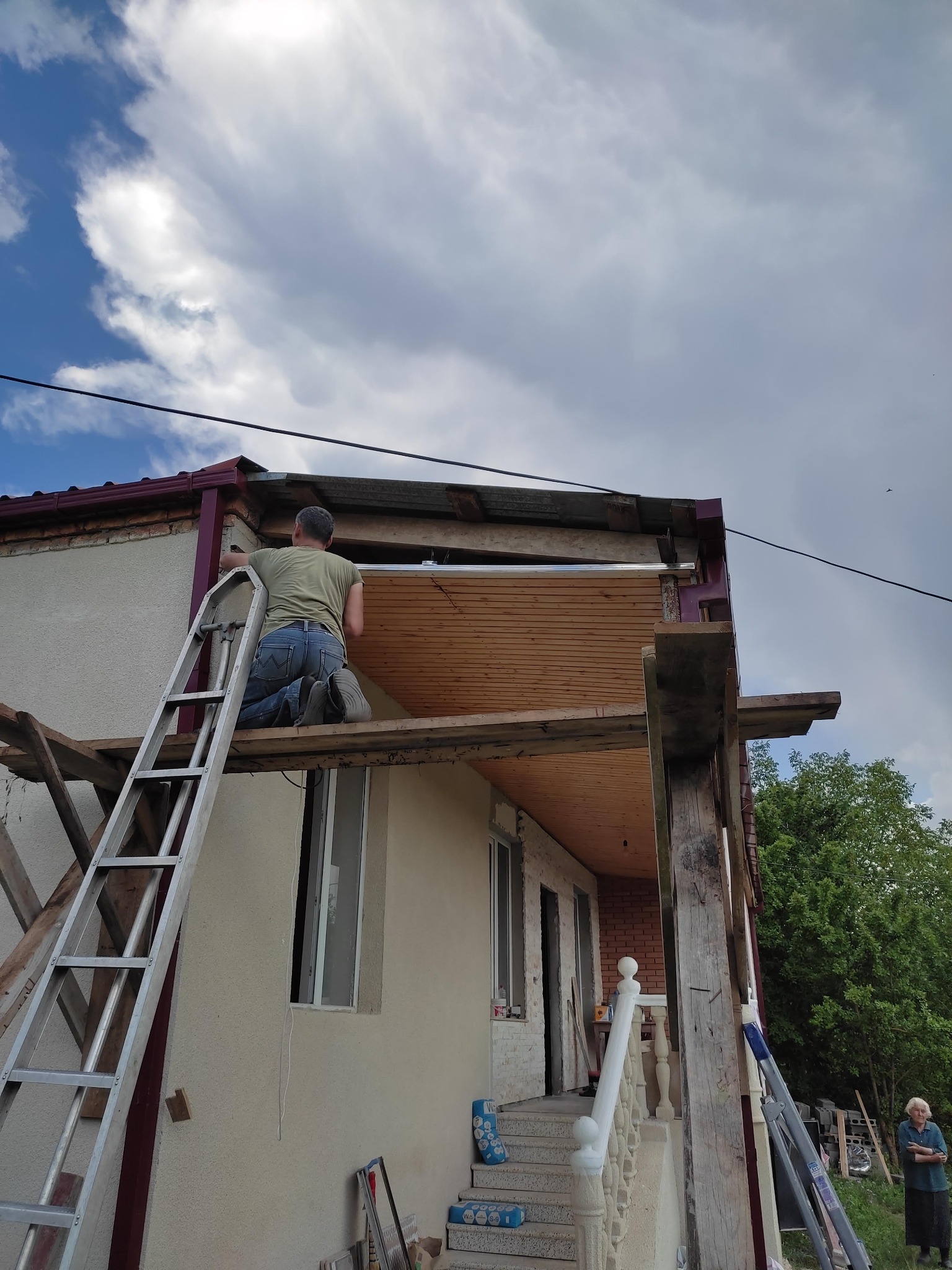 Pracownik na kolanach na rusztowaniu, montuje drewniane deski pod zadaszeniem domu jednorodzinnego. Widoczna drabina oparta o ścianę budynku, obok stoi starsza kobieta.