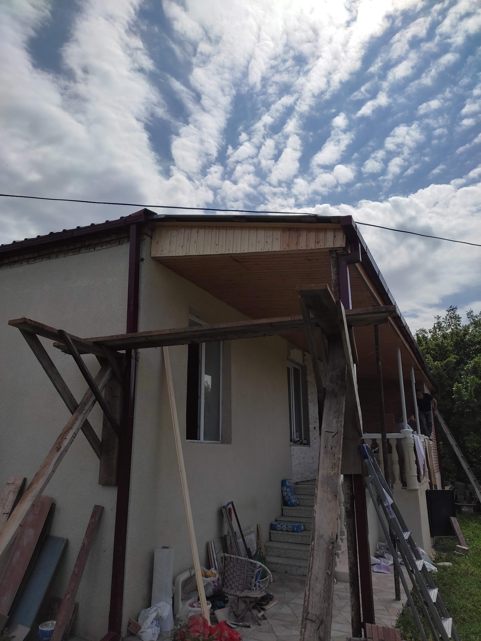 Dom w trakcie prac wykończeniowych werandy, widoczne drewniane deski sufitowe, rusztowanie i drabina oparta o ścianę, na pierwszym planie porozrzucane materiały budowlane.