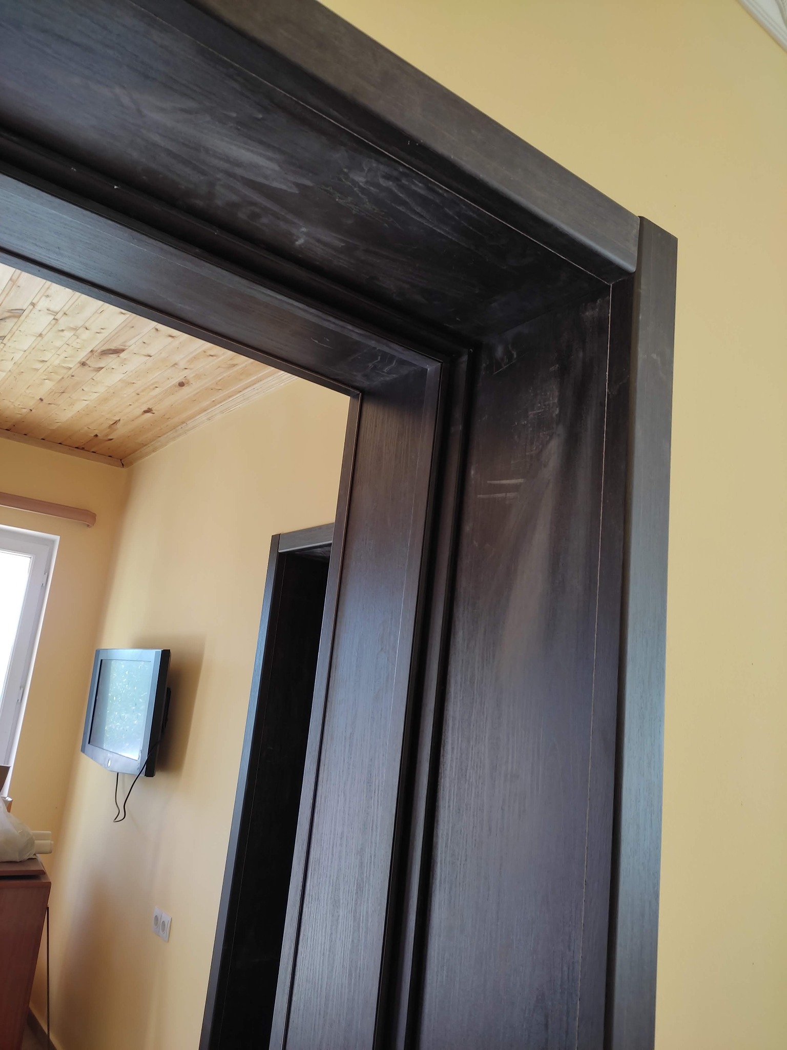 Ciemna, drewniana ościeżnica drzwiowa z widocznymi śladami użytkowania i drobnymi zarysowaniami, na tle żółtej ściany i drewnianego sufitu.