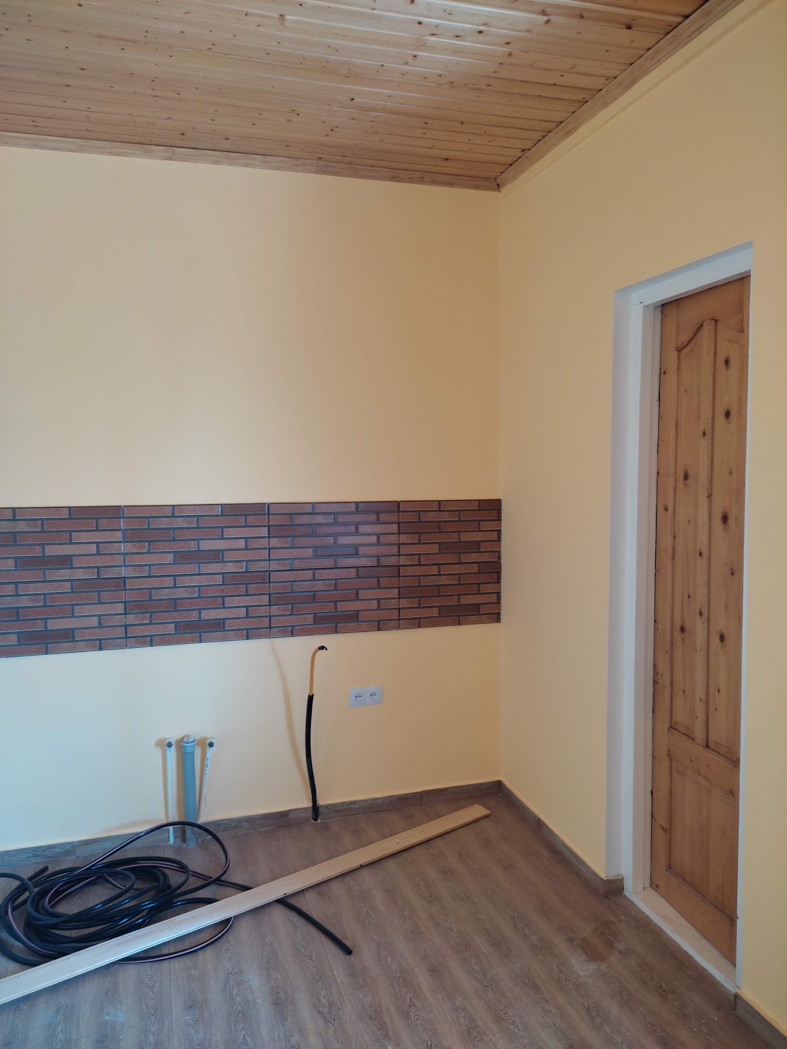 Pokój w trakcie prac: częściowo ułożona dekoracyjna okładzina z cegły, drewniany sufit, widoczne przewody elektryczne i rury hydrauliczne, drewniane panele podłogowe i ościeżnica drewnianych drzwi.