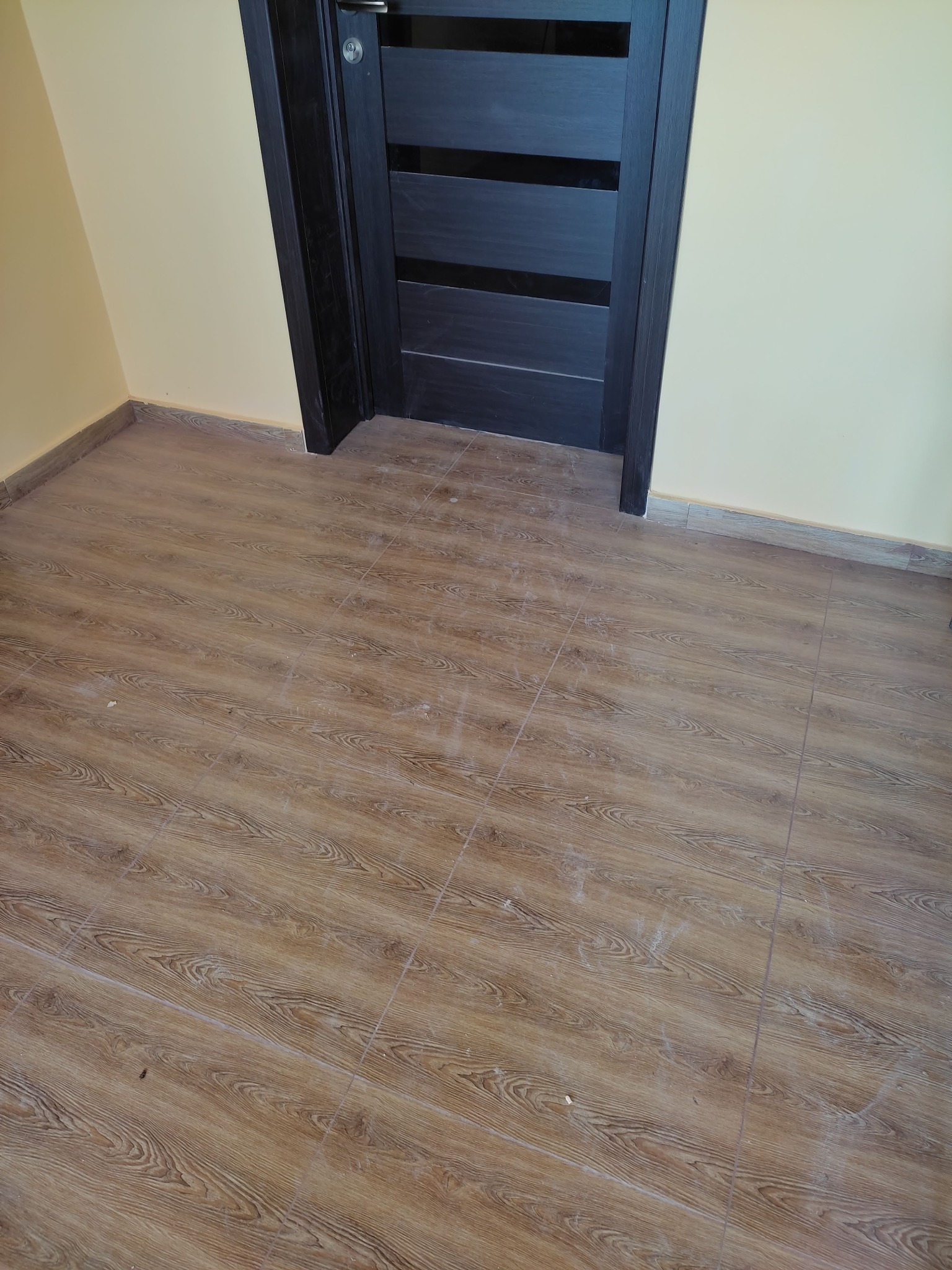 Ułożone panele podłogowe z wzorem drewna, widoczny fragment ściany w kolorze kremowym i ciemne, nowoczesne drzwi z poziomymi wstawkami.