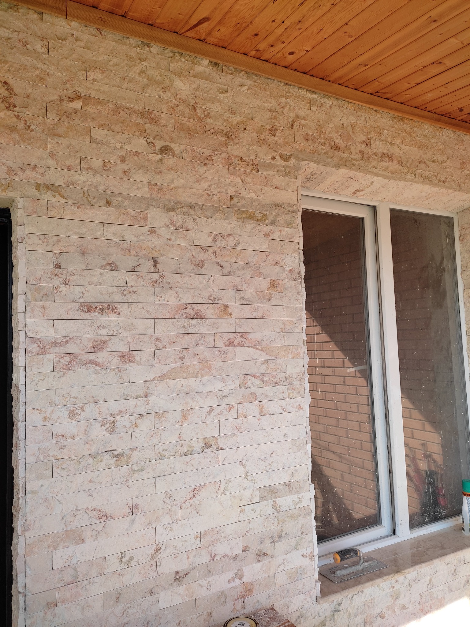 Ściana zewnętrzna wyłożona jasnymi płytkami imitującymi kamień, widoczne okno z białymi ramami, drewniane wykończenie sufitu oraz narzędzia glazurnicze na parapecie.