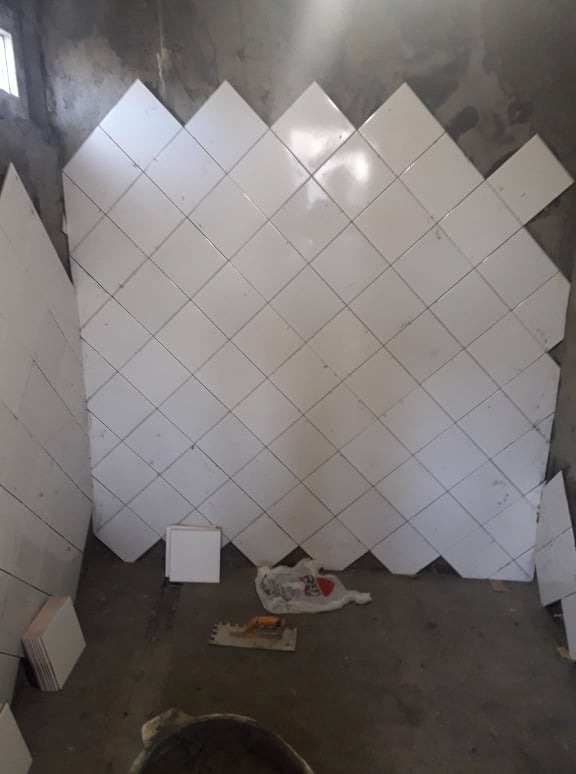 Ściana w trakcie układania białych, kwadratowych płytek diagonalnie, widoczne niedociągnięcia przy krawędziach, zapasowe płytki na podłodze oraz narzędzia glazurnicze i wiadro.