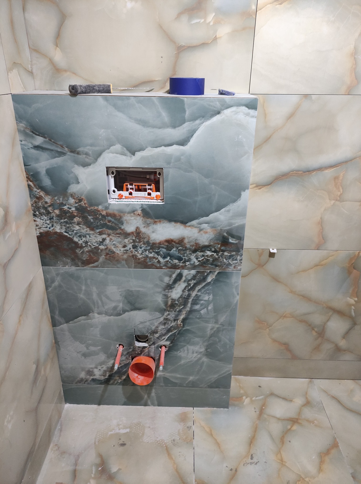 Montaż instalacji podtynkowej WC na tle marmurowych płytek w odcieniach zieleni i beżu, widoczne wystające rury kanalizacyjne i narzędzia glazurnicze na górnej krawędzi ściany.