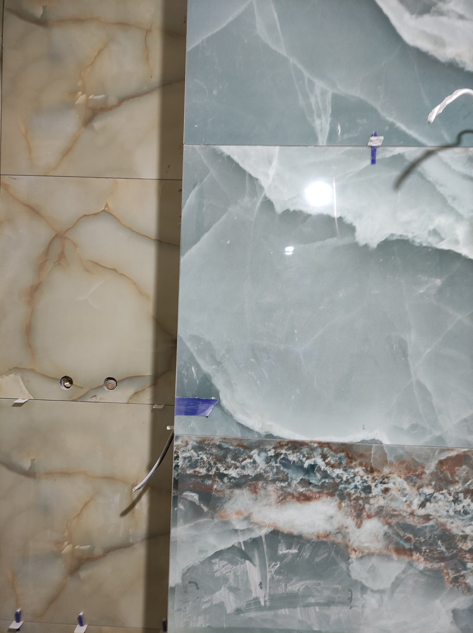 Ułożone płytki imitujące marmur w odcieniach beżu i błękitu, widoczne plastikowe kliny do poziomowania, przygotowane otwory pod instalację.