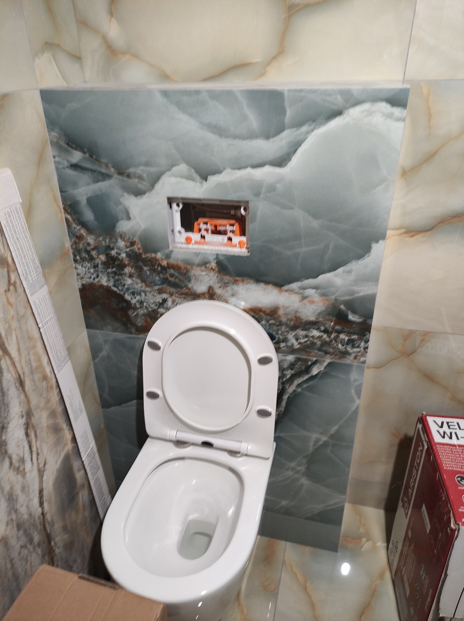 Zamontowana, biała toaleta wisząca na tle ściany wyłożonej płytkami imitującymi marmur, widoczna instalacja podtynkowa.